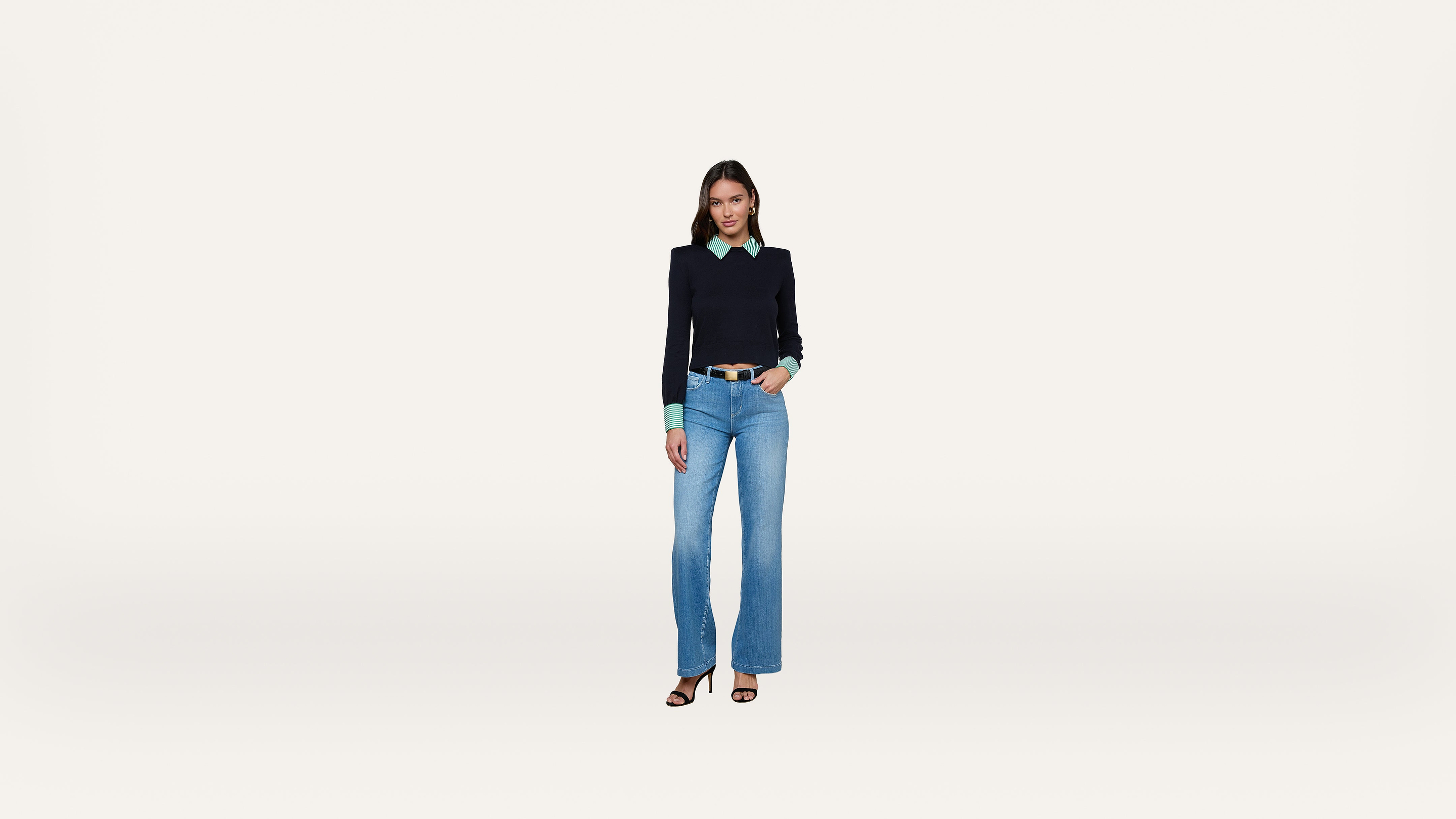 A woman stands facing forward, wearing a black sweater over a green shirt, blue jeans, and black heeled sandals, against a plain white background.