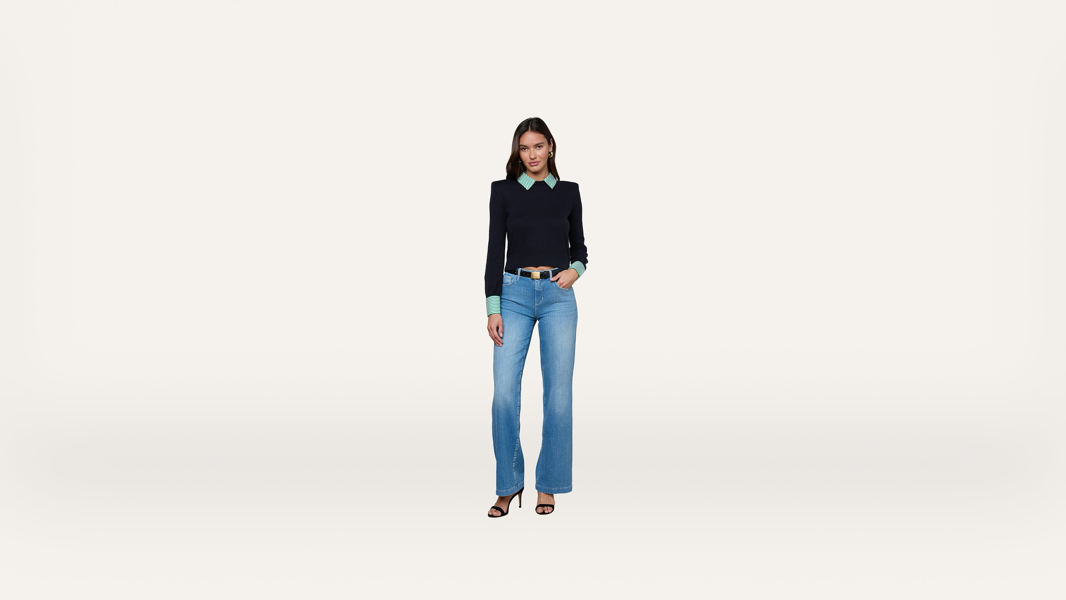 A woman stands facing forward, wearing a black sweater over a green shirt, blue jeans, and black heeled sandals, against a plain white background.