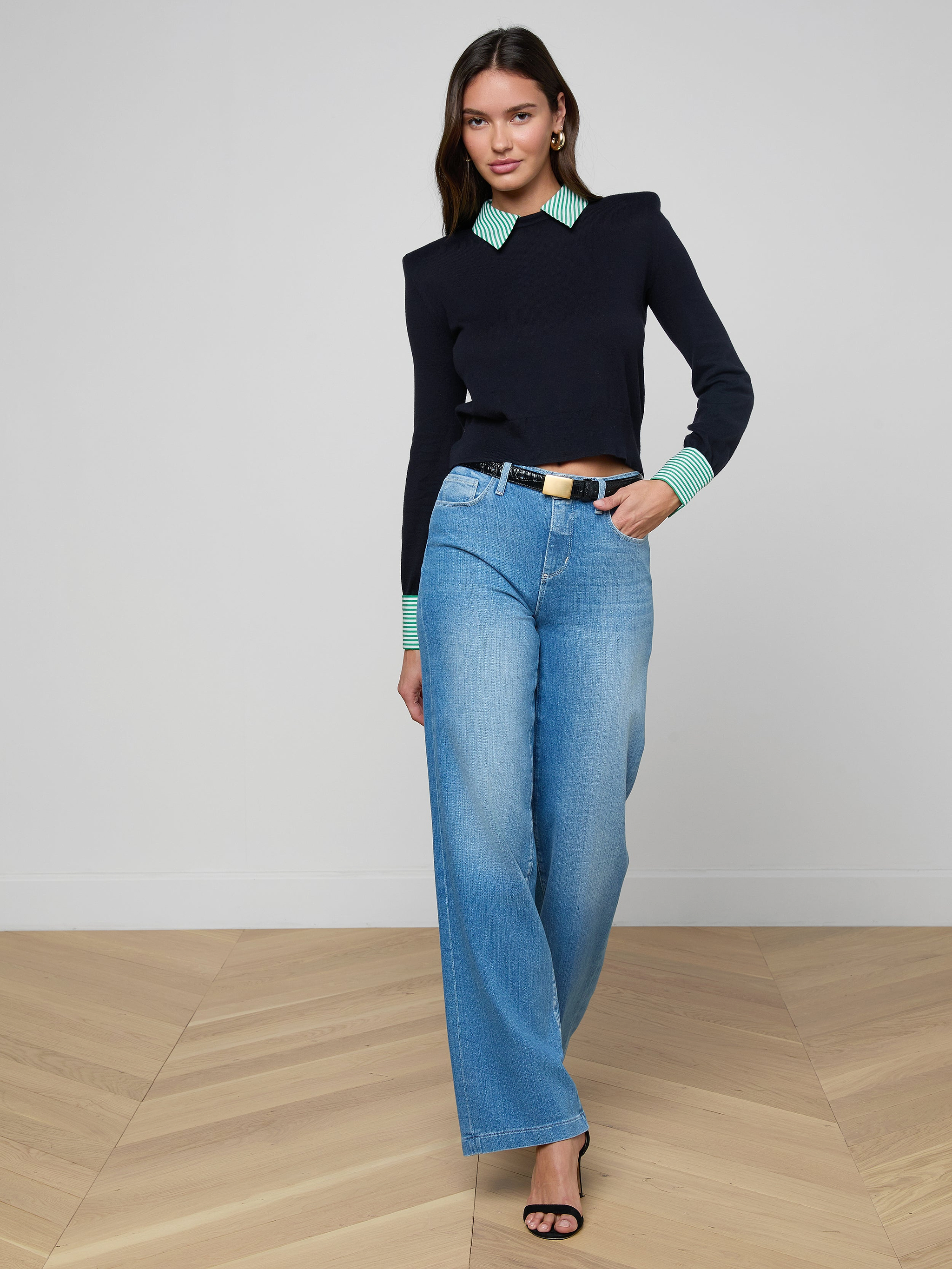 A woman with long brown hair and a neutral expression stands on a wooden floor wearing the April Poplin Collar Pullover—navy blue with a green striped collar—paired with light blue wide-leg jeans, black belt and heeled sandals, and gold earrings.