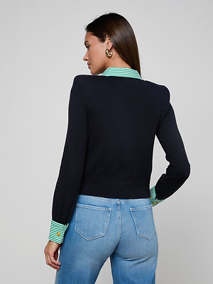 A woman with brown hair wears the April Poplin Collar Pullover—a black featherweight knit with green and white striped collar and cuffs—paired with light blue jeans and hoop earrings, standing against a plain light background.