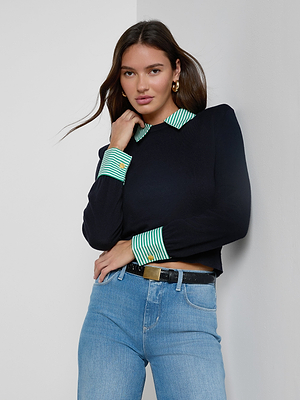 A woman with long brown hair wears the April Poplin Collar Pullover with a striped collar and green and white cuffs, paired with blue jeans and a black belt, standing against a white wall.
