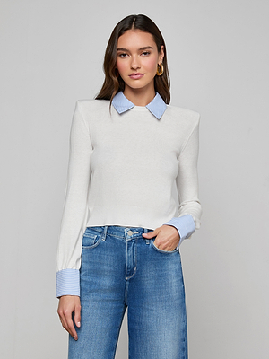 A woman poses against a plain backdrop in the April Poplin Collar Pullover, featuring a featherweight knit with blue striped collar and cuffs, paired with high-waisted blue jeans. She has long brown hair, gold hoop earrings, and one hand in her pocket.