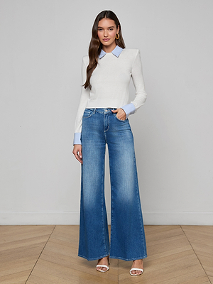 A woman with long brown hair poses indoors against a plain background, wearing the April Poplin Collar Pullover—a featherweight knit white long-sleeve top with a blue striped collar—paired with high-waisted wide-leg jeans and white heeled sandals.