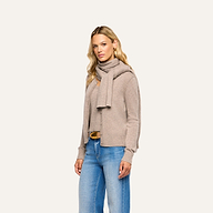 A person wears a pebbled tan Palais Cashmere Scarf draped over a matching cardigan and top with blue jeans.