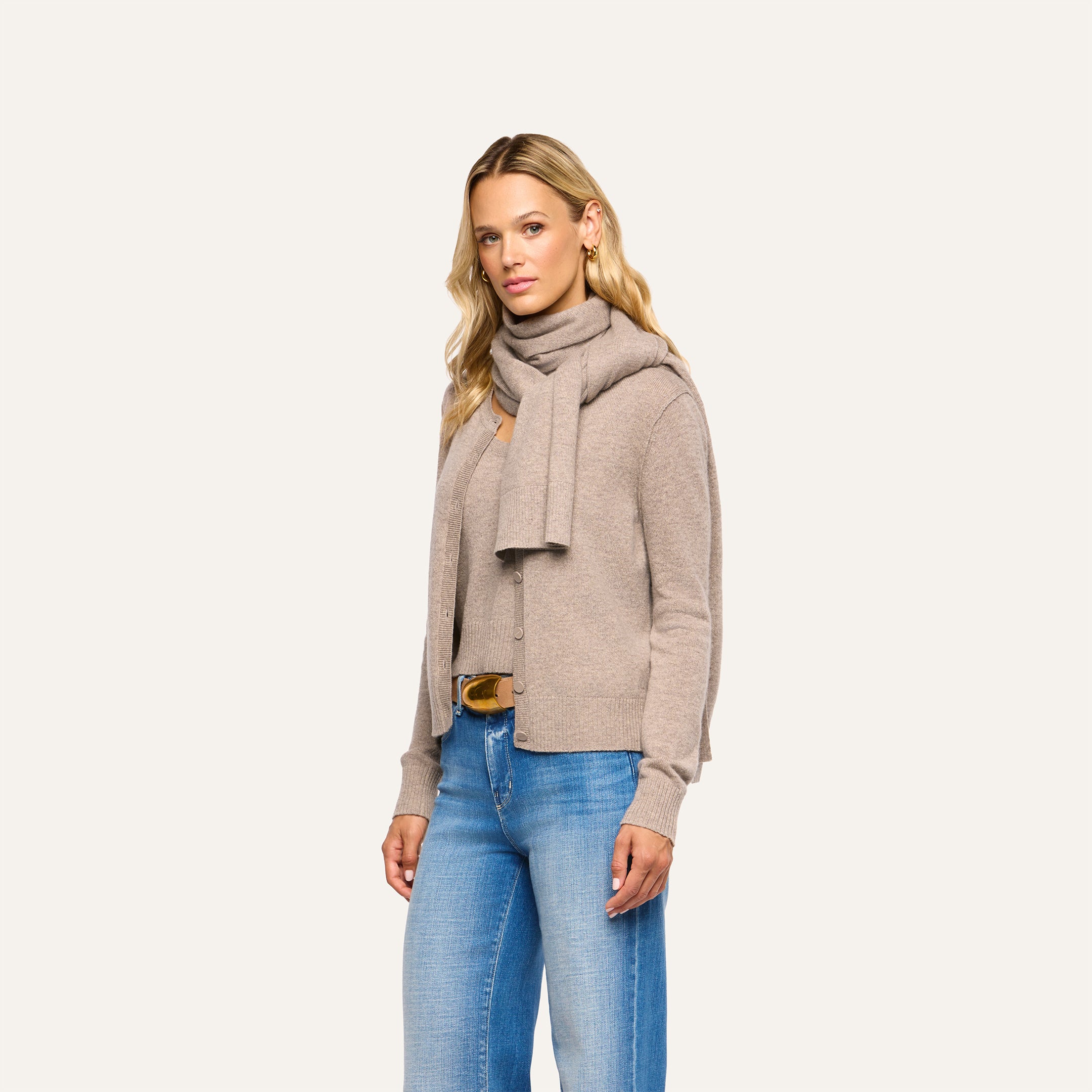 A person wears a pebbled tan Palais Cashmere Scarf draped over a matching cardigan and top with blue jeans.