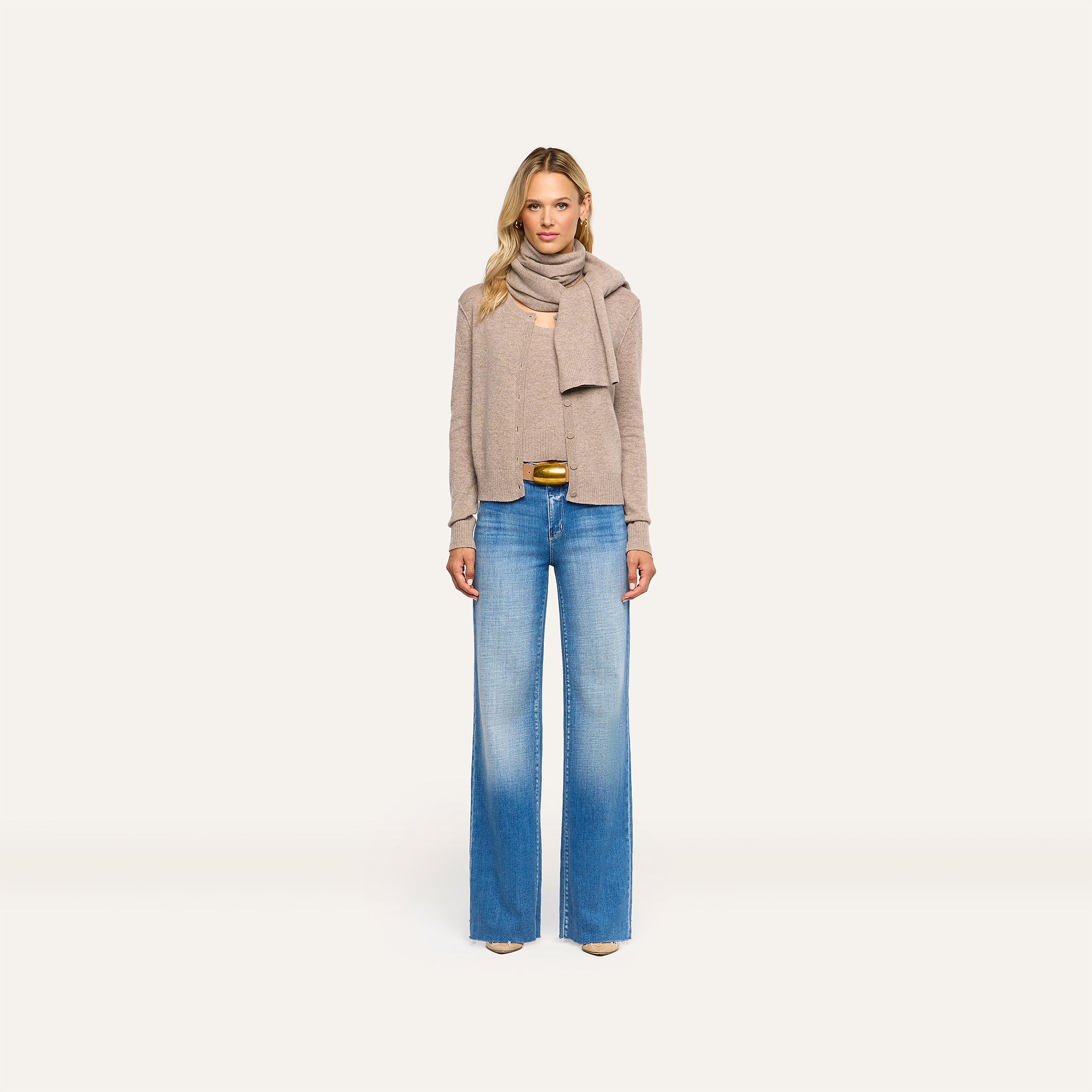 A woman wearing a tan Palais Cashmere Scarf draped over a matching cardigan and tank top with blue wide-leg jeans.