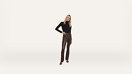 A woman with long blonde hair, wearing a black long-sleeve top, brown leather pants, and black heeled sandals, stands against a plain white background.
