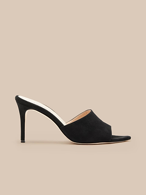 The Lolita Suede Open-Toe Mule is a black, minimalist high stiletto heel in suede, featuring an open-toe design and shown in profile against a beige background.