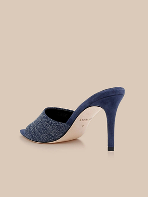 The Lolita Denim Open-Toe Mule features a blue denim upper and slim blue suede heel, shown against a beige background.