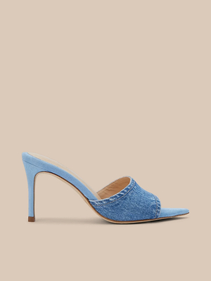 The Lolita Denim Open-Toe Mule is a single light blue mule sandal with a denim upper, slim stiletto heel, and open toe, shown on a beige background.