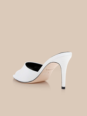 The Lolita Leather Open-Toe Mule showcases white lizard-embossed leather, a thin high heel, and a minimalist peep-toe design. Displayed angled on a light beige background, it features a black interior and an elegantly textured surface.
