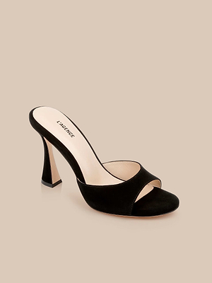 The Avery Suede Open-Toe Mule, a black sculpted stiletto with a flared heel and nude insole printed with LAGENCE, is displayed on a beige background.