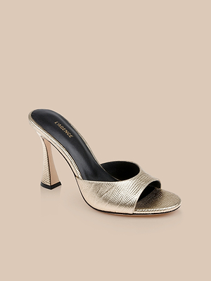 The Avery Leather Open-Toe Mule features gold textured leather, a sculpted stiletto mule silhouette, open toe, black insole, and a flared heel, displayed on a beige background.