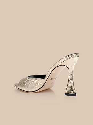 The Avery Leather Open-Toe Mule, featuring gold leather and a sculpted flared heel, is shown against a beige background, highlighting the elegance of this stylish stiletto mule.