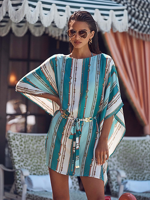 A woman wearing sunglasses and the Alia Cover-Up Tunic stands confidently outdoors in front of a building with a striped awning and patterned furniture.