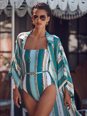 A woman with wet, slicked-back hair and gold hoop earrings walks outdoors under a fringed umbrella, wearing sunglasses, a high-leg cut sage Tory Bandeau One-Piece Swimsuit by L'AGENCE and a matching cover-up.