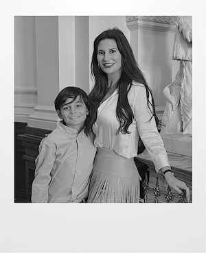A woman and a young boy stand close together indoors, smiling at the camera. The woman wears a long-sleeved blouse and a pleated skirt, while the boy wears a button-up shirt. Architectural details and a statue are visible behind them.