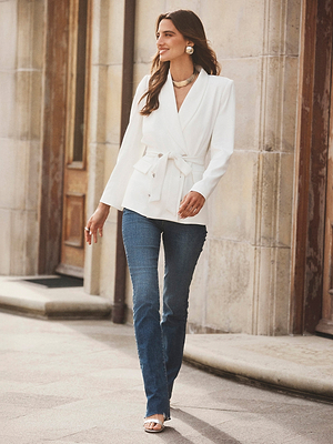A woman with long brown hair walks outdoors by a stone building, smiling slightly and wearing the Aven Wrap Blazer in white, blue jeans, white sandals, statement earrings, and layered necklaces.