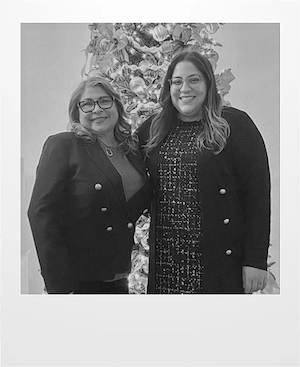 Two women smiling and standing side by side in front of a decorated Christmas tree. Both are wearing glasses and dark-colored jackets, and the photo is in black and white.