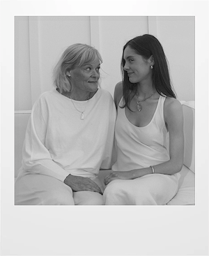 An older woman and a younger woman sit closely together on a couch, facing each other and smiling warmly. Both are dressed in light-colored clothing in a softly lit, minimalist setting.