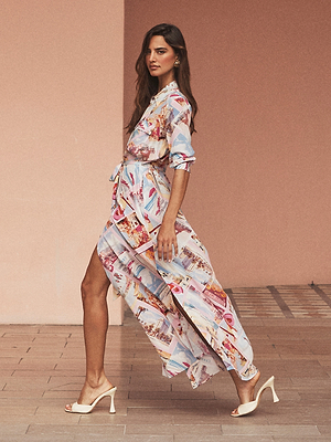 A woman confidently walks in heeled sandals, wearing the Cameron Silk Shirt Dress with a vibrant postcard print and high slit, set against a peach wall and tiled floor.