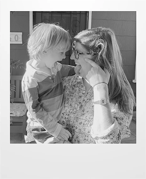 A woman wearing glasses smiles and holds a young child in her arms. They are touching foreheads and looking at each other happily, outdoors in natural light.