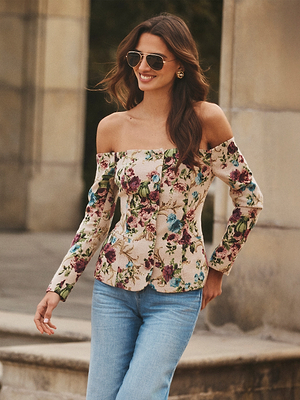A woman wearing sunglasses and the Alice Blazer Top over her off-the-shoulder long sleeve top with blue jeans smiles while walking outdoors near stone columns.