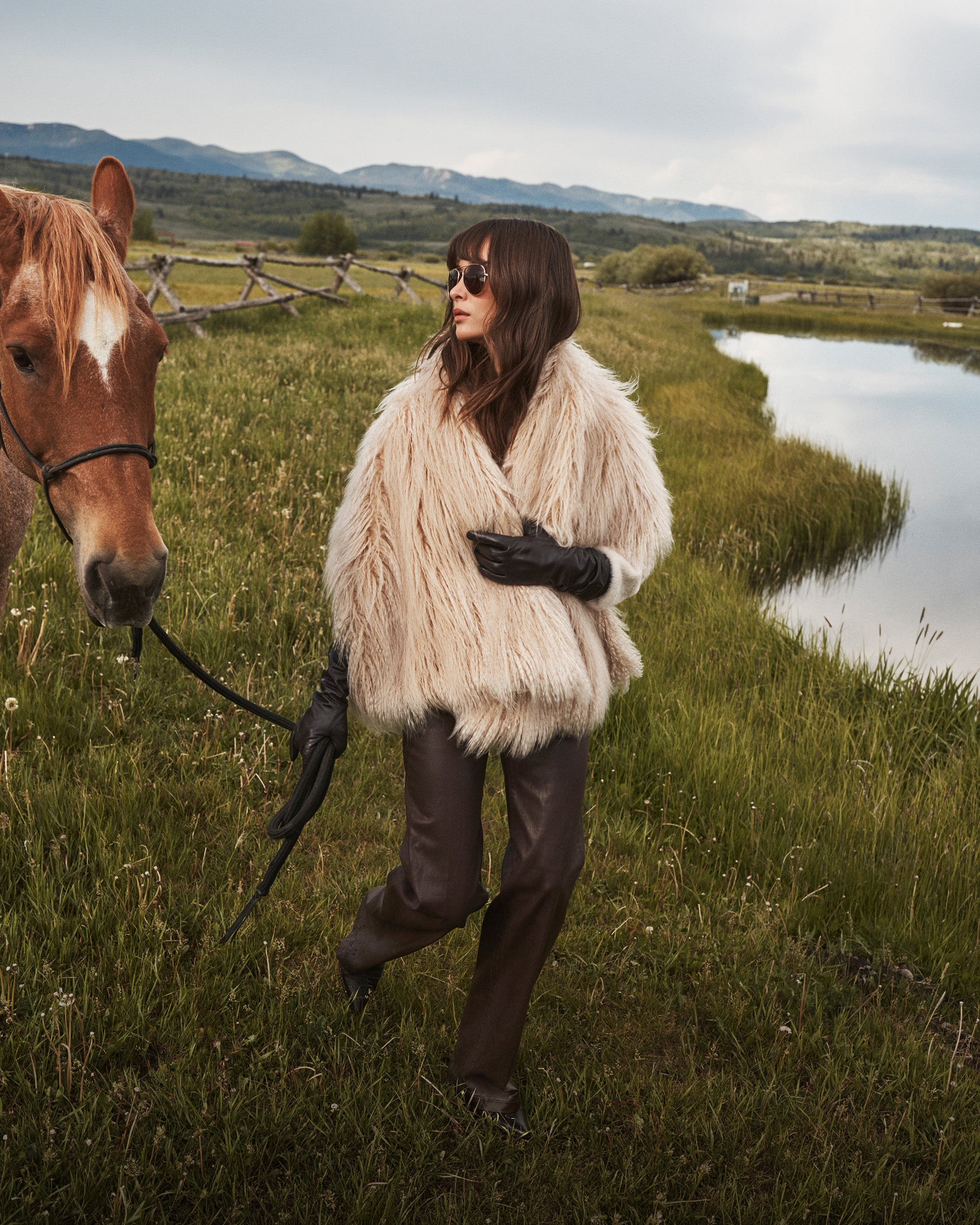 A woman in a shaggy fur coat, black gloves, and sunglasses stands on grass, holding a horses reins near a pond, with mountains in the background.
