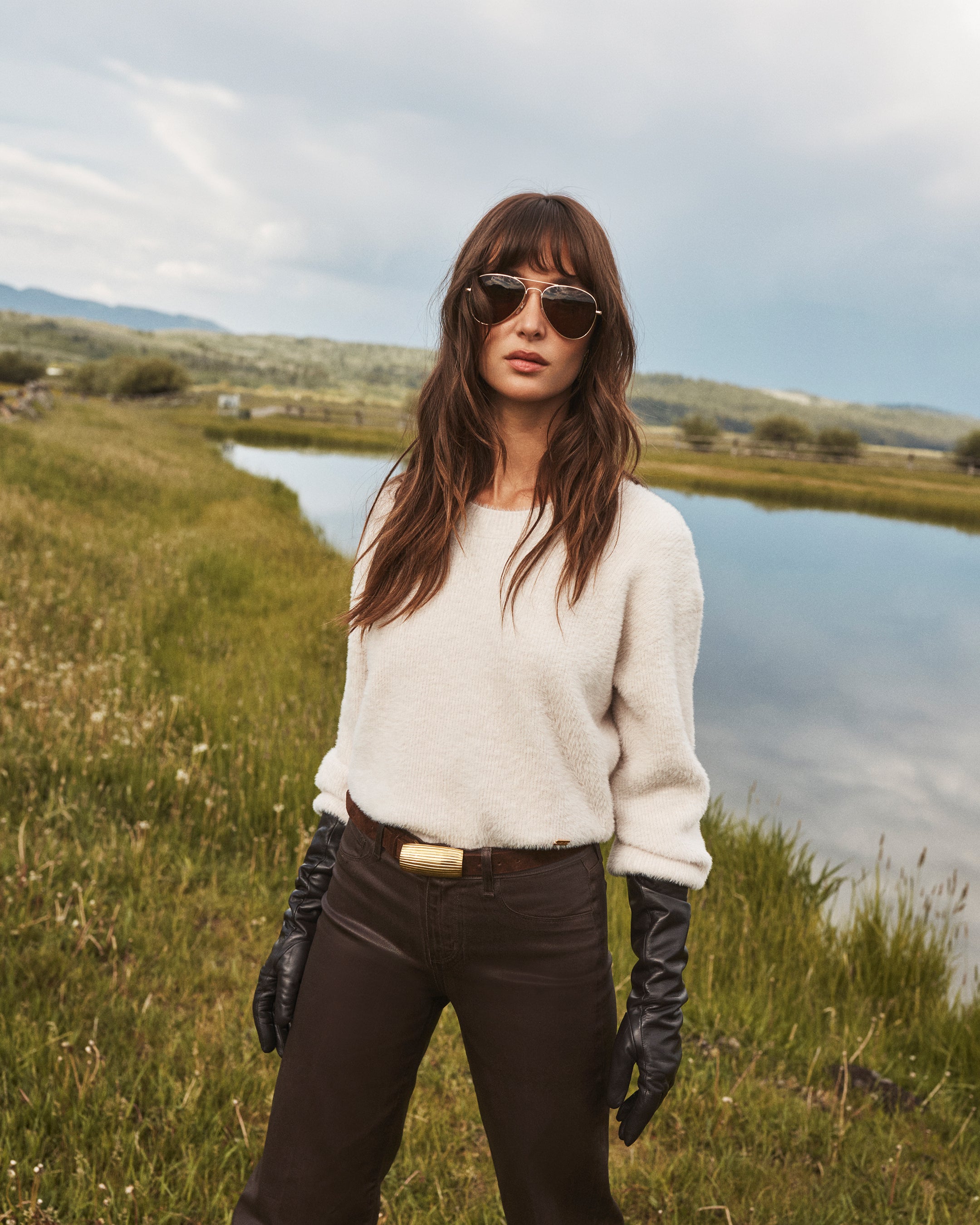 A woman wearing sunglasses, a white sweater, brown pants, and black gloves stands by a grassy pond on a cloudy day.