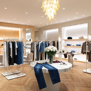 A modern clothing boutique interior with multiple clothing racks, a marble display table, wood flooring, and a decorative gold chandelier. A pair of blue jeans is draped over the front of the round marble table, and a vase of white flowers sits in the center.