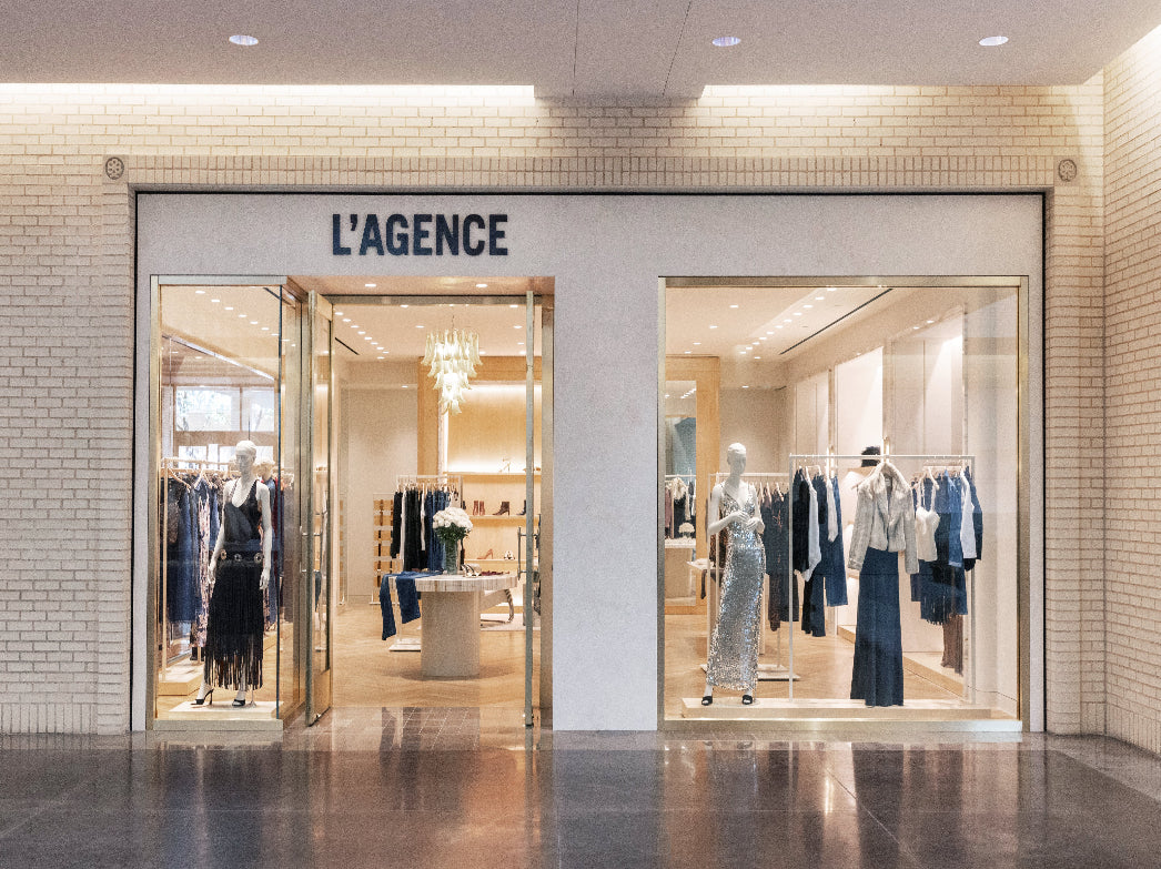 L'Agence clothing boutique storefront with mannequins and racks of apparel visible through large glass windows, set against a beige brick wall and polished floor.