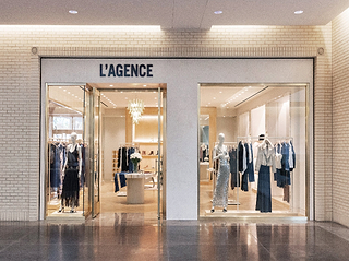 L'Agence clothing boutique storefront with mannequins and racks of apparel visible through large glass windows, set against a beige brick wall and polished floor.