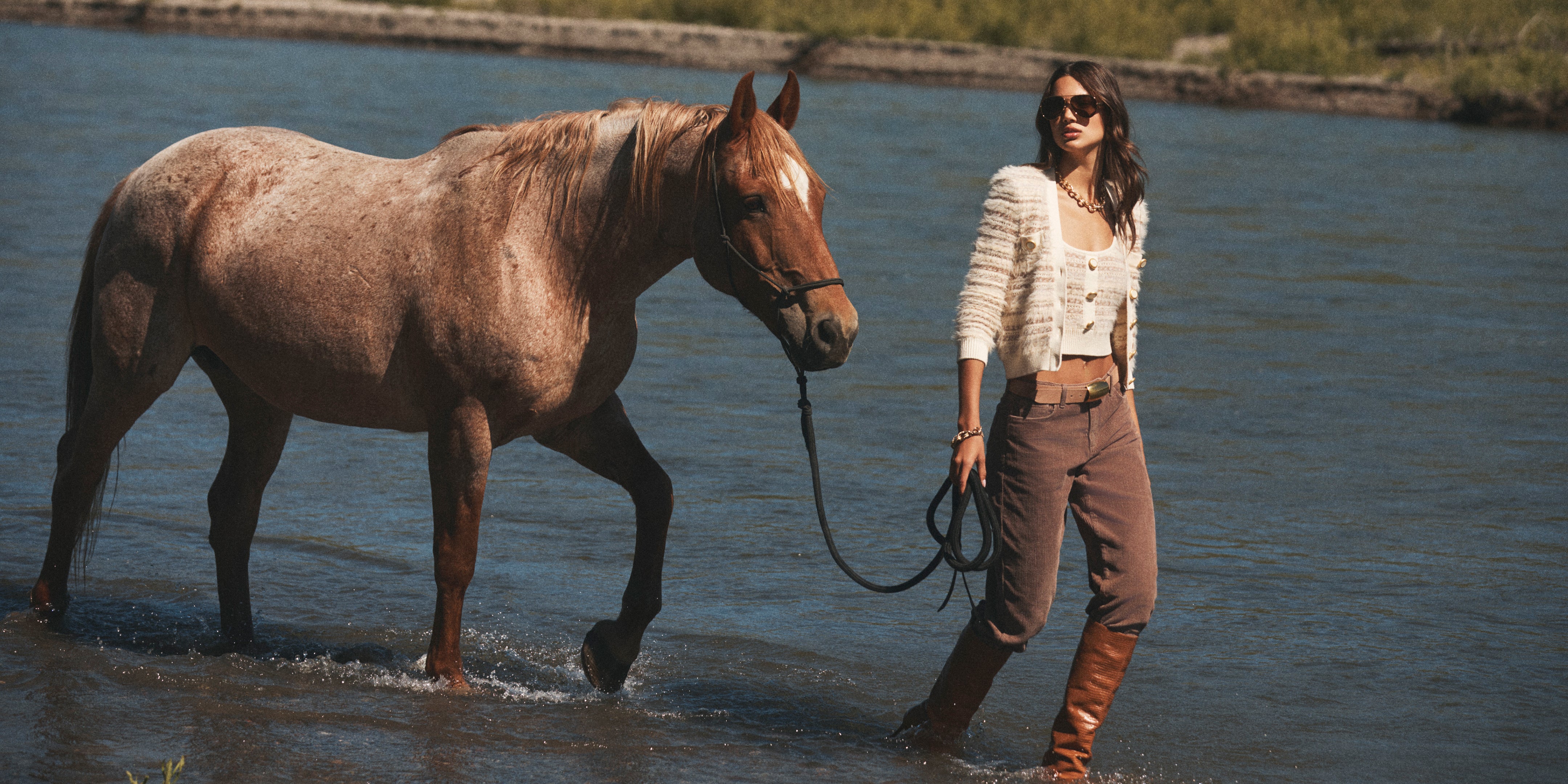 A woman wearing sunglasses and boots leads a brown horse on a leash through a shallow river, with greenery visible in the background on a sunny day.