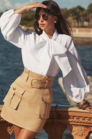 A woman wearing sunglasses, a white blouse with a large bow, and a beige belted skirt stands by a waterfront, shielding her eyes from the sun with one hand and resting her other hand on a stone railing.