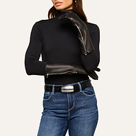 A woman wearing a black turtleneck, blue jeans, a wide black belt with a large silver buckle, and long luxury Carmen Gloves in lambskin, poses with one hand on her neck and the other arm across her body.