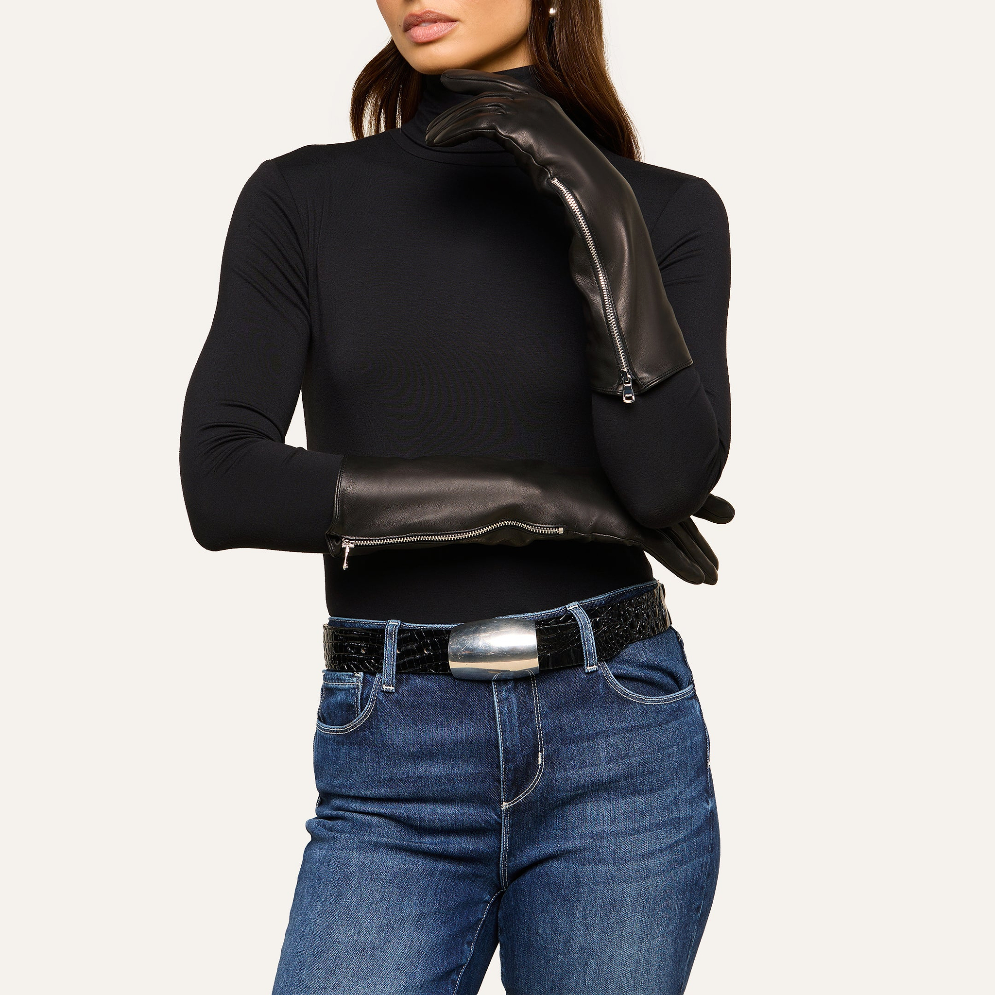 A woman wearing a black turtleneck, blue jeans, a wide black belt with a large silver buckle, and long luxury Carmen Gloves in lambskin, poses with one hand on her neck and the other arm across her body.