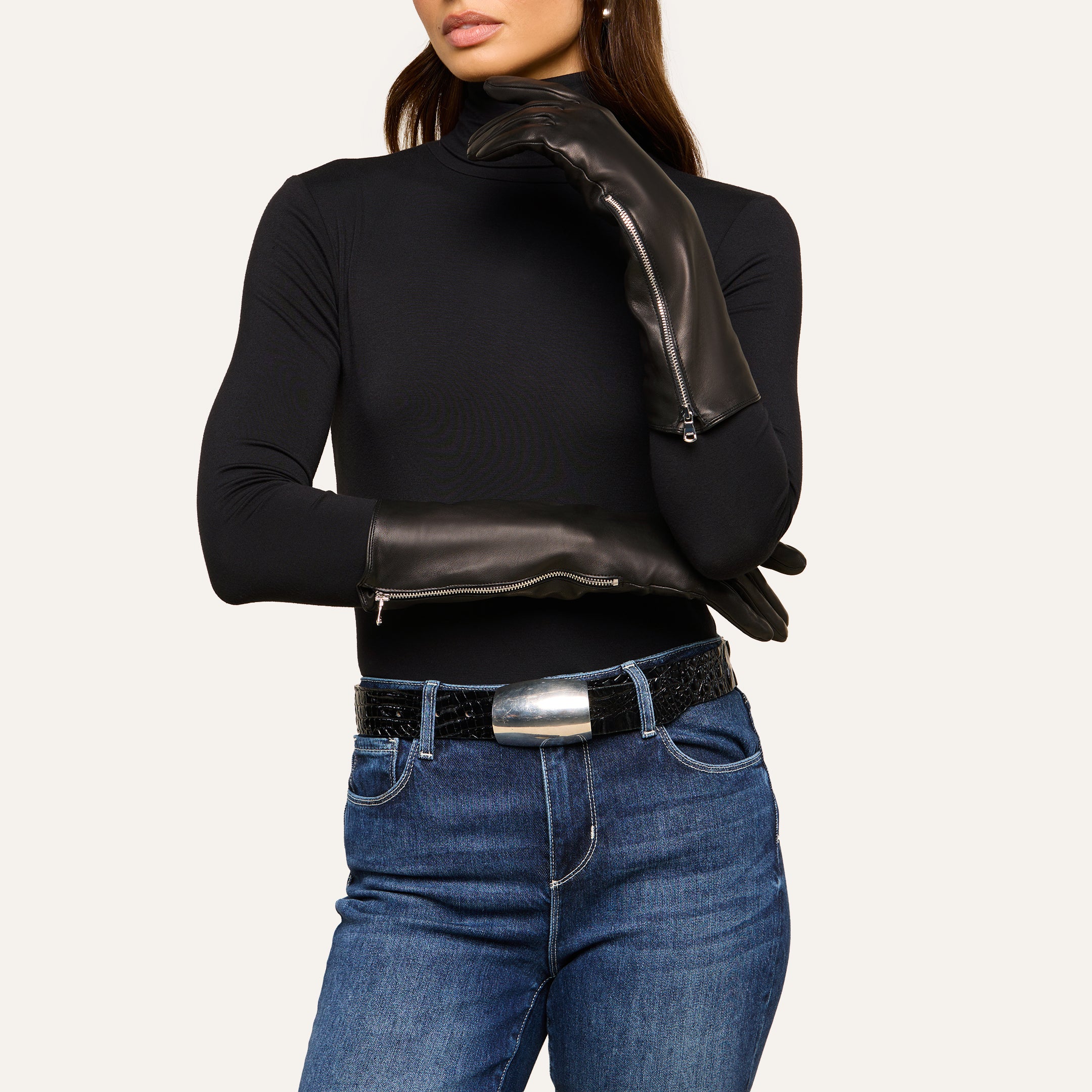 A woman wearing a black turtleneck, blue jeans, a wide black belt with a large silver buckle, and long luxury Carmen Gloves in lambskin, poses with one hand on her neck and the other arm across her body.