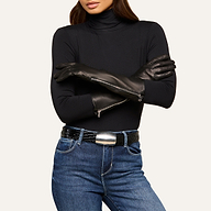 A person with their arms crossed is wearing the Carmen Gloves—luxury lambskin gloves with zippers—alongside a black turtleneck, blue jeans, and a black textured belt with a large silver buckle. Their face is not fully visible.