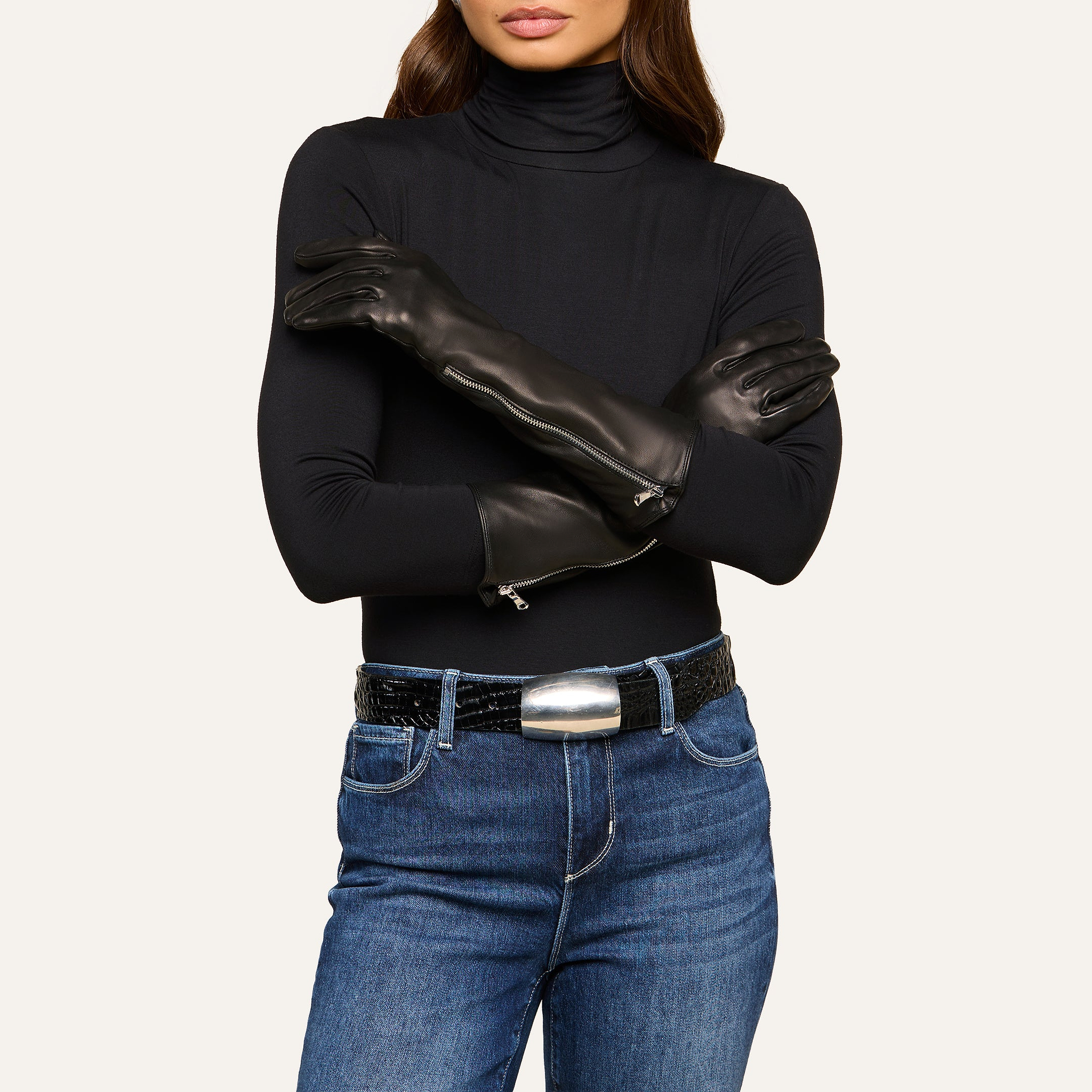 A woman wearing a black turtleneck, blue jeans, and a black belt with a silver buckle stands with her arms crossed, showcasing the Carmen Gloves—luxurious long black leather gloves featuring zippers.