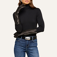 Wearing the Carmen Gloves—long, luxury leather gloves—a woman poses with one arm across her body. She is also dressed in a black turtleneck, blue jeans, and a silver-buckled belt, with her face partially out of frame.