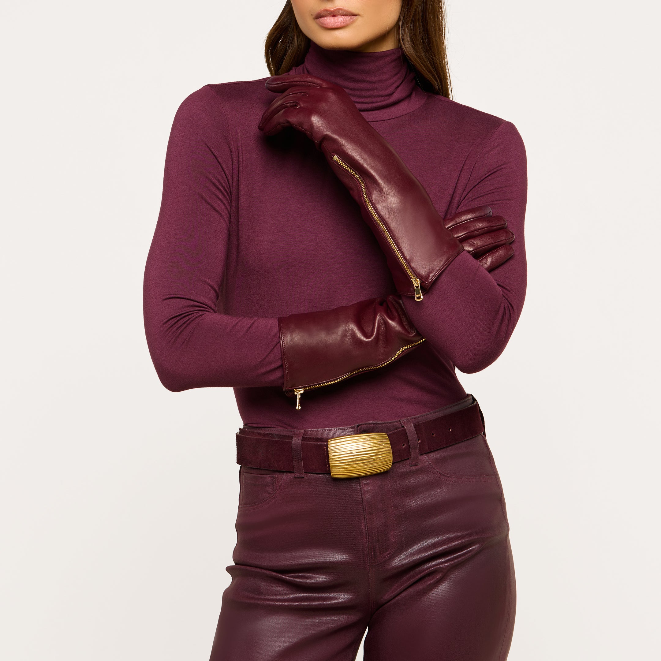Wearing the Carmen Gloves—long luxury lambskin leather gloves featuring zippers—a person dressed in a maroon turtleneck, matching leather pants, and a thick belt poses with arms crossed against a plain background.