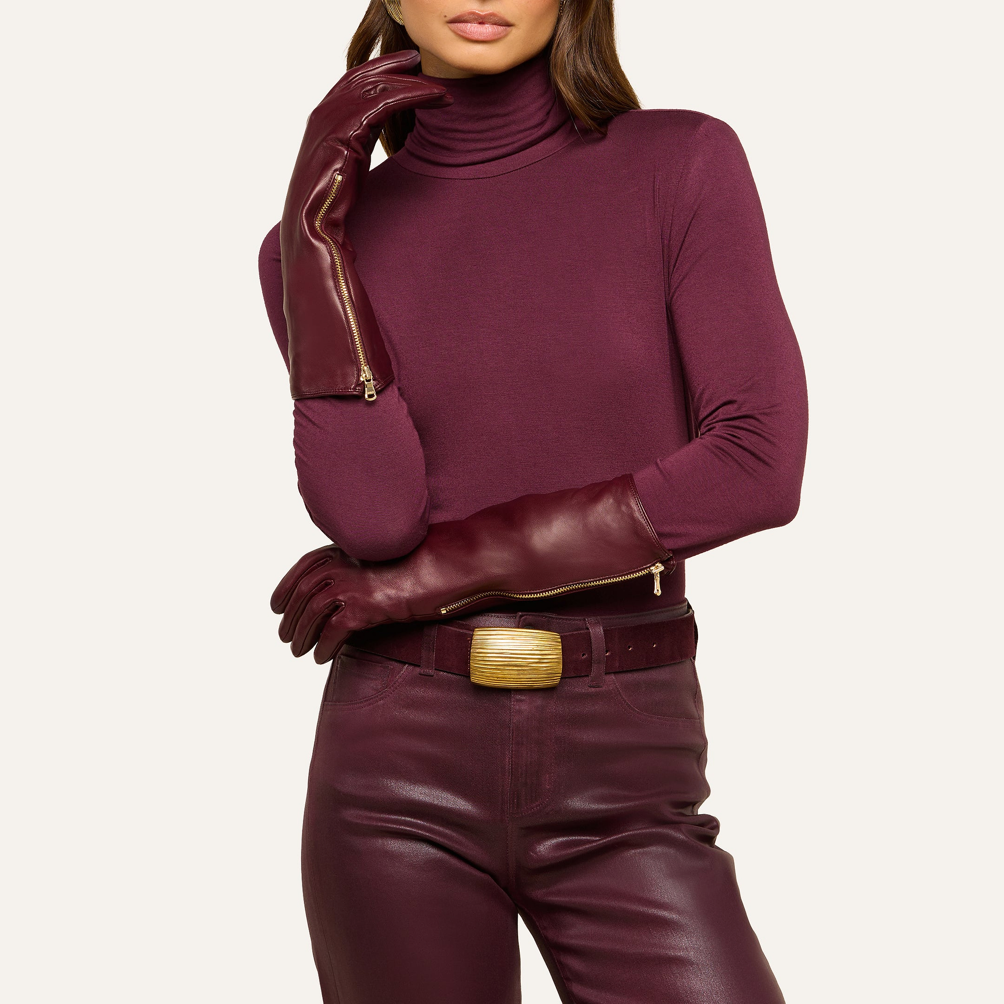 A person wearing a burgundy turtleneck, matching Carmen Gloves crafted from lambskin leather, and pants with a wide beige belt poses with one hand on their face and the other resting on their arm.