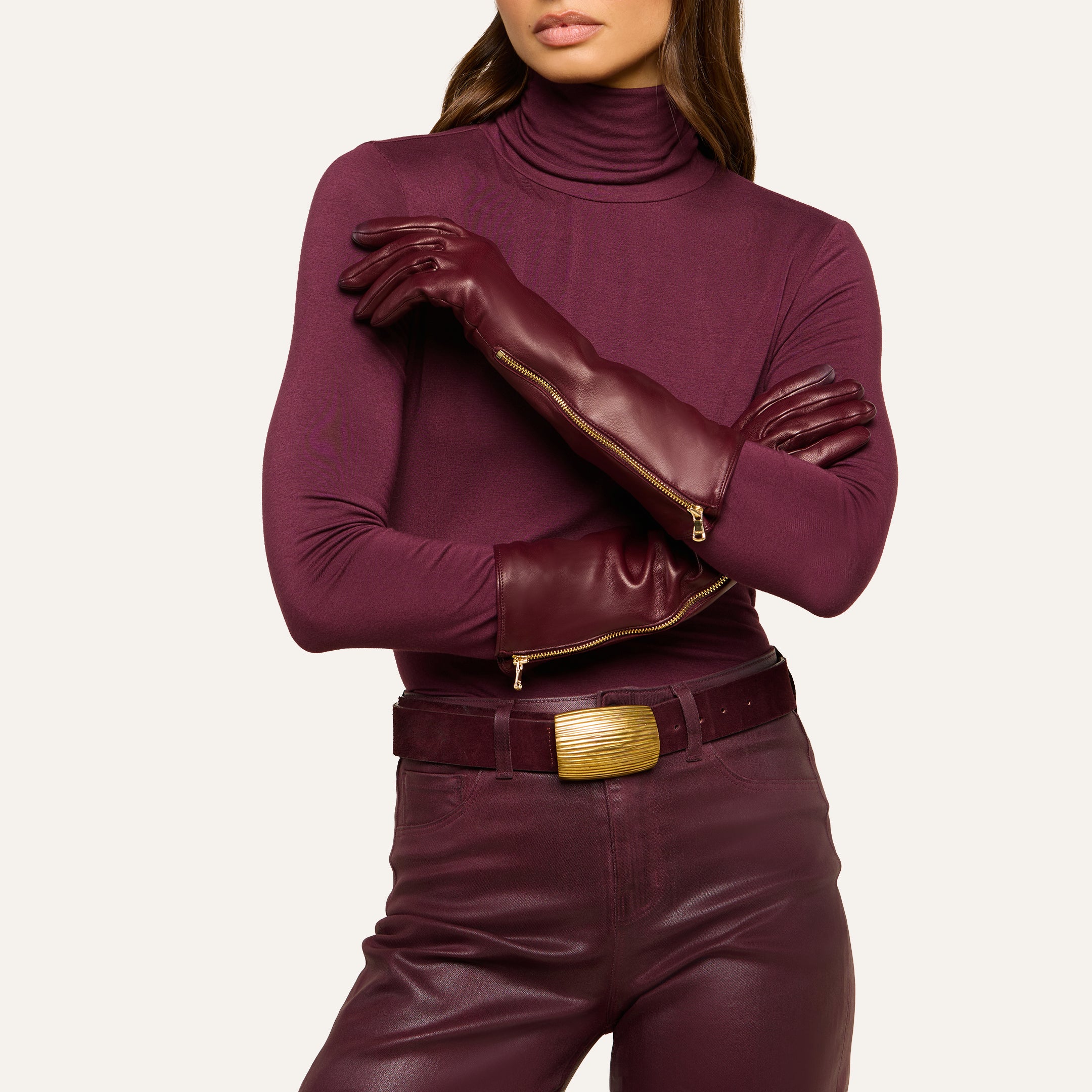 A woman stands with arms crossed, wearing a maroon turtleneck, high-waisted maroon pants with a wide belt and gold buckle, and the Carmen Gloves—luxury lambskin leather gloves. Her face is partially out of frame.