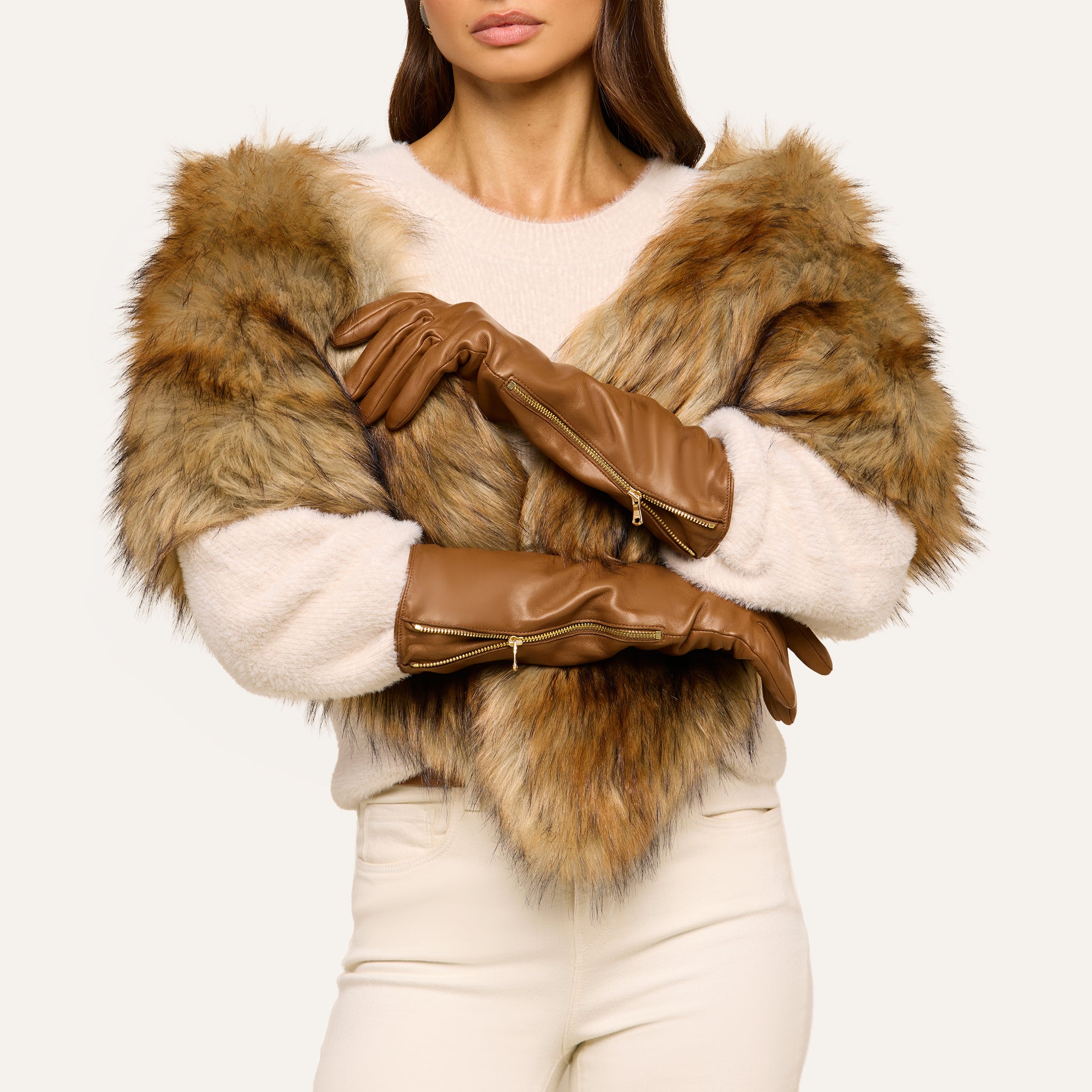 A woman stands with arms crossed against a plain background, partially revealing her face. She is wearing a beige sweater, white pants, a faux fur shawl, and the Carmen Gloves—long brown silk-lined gloves.