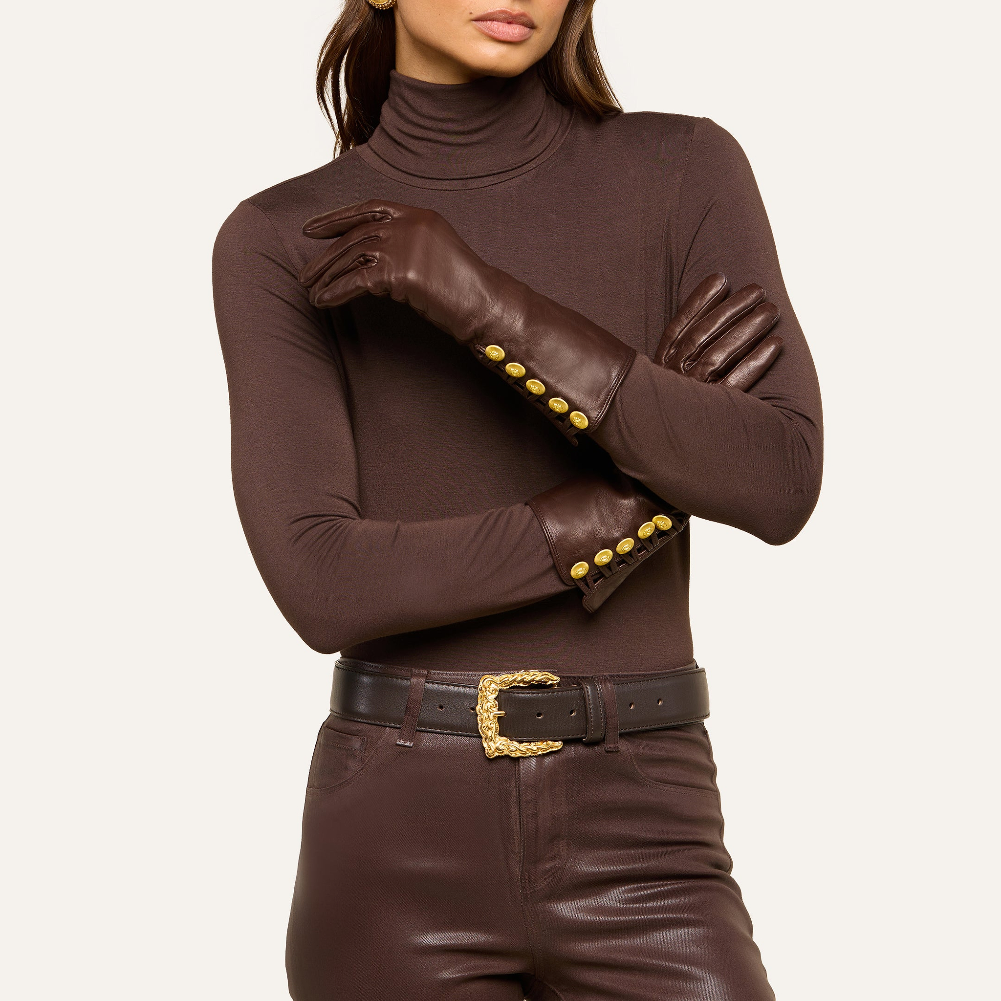A woman stands with her arms crossed, wearing a brown turtleneck, stylish Tosca Gloves featuring gold buttons, coordinated brown pants, and a black belt with a gold buckle.