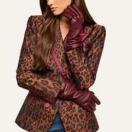 A woman with long brown hair is pictured wearing a leopard print blazer accented with burgundy trim and buttons, her lower face visible as she poses with one arm crossed and the other hand—elegantly gloved in Boheme Gloves crafted from luxury burgundy lambskin leather—resting thoughtfully beneath her chin.
