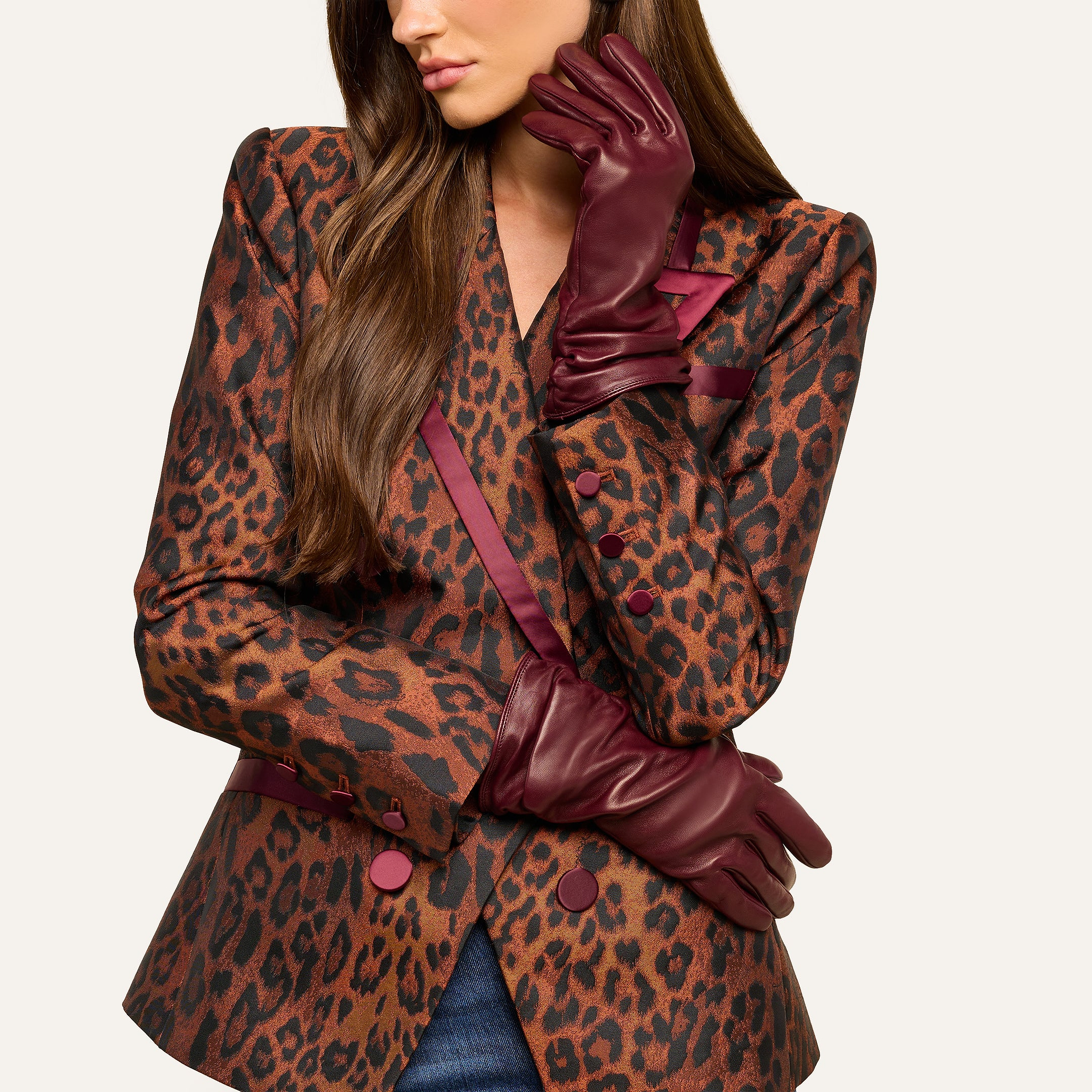 A woman with long brown hair is pictured wearing a leopard print blazer accented with burgundy trim and buttons, her lower face visible as she poses with one arm crossed and the other hand—elegantly gloved in Boheme Gloves crafted from luxury burgundy lambskin leather—resting thoughtfully beneath her chin.