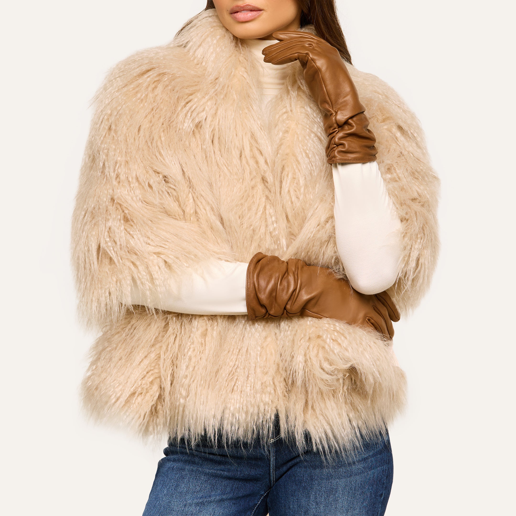 Woman wearing a fluffy beige fur coat, a white turtleneck, blue jeans, and elegant Boheme Gloves crafted from lambskin. Her head is slightly tilted, and her face is partially visible.