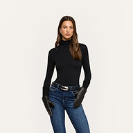 A woman with long brown hair wearing a black turtleneck, blue jeans, a belt, and earrings stands confidently against a plain light background. She completes her elegant ensemble with Tempo Mittens, adding a touch of sophistication to her look.