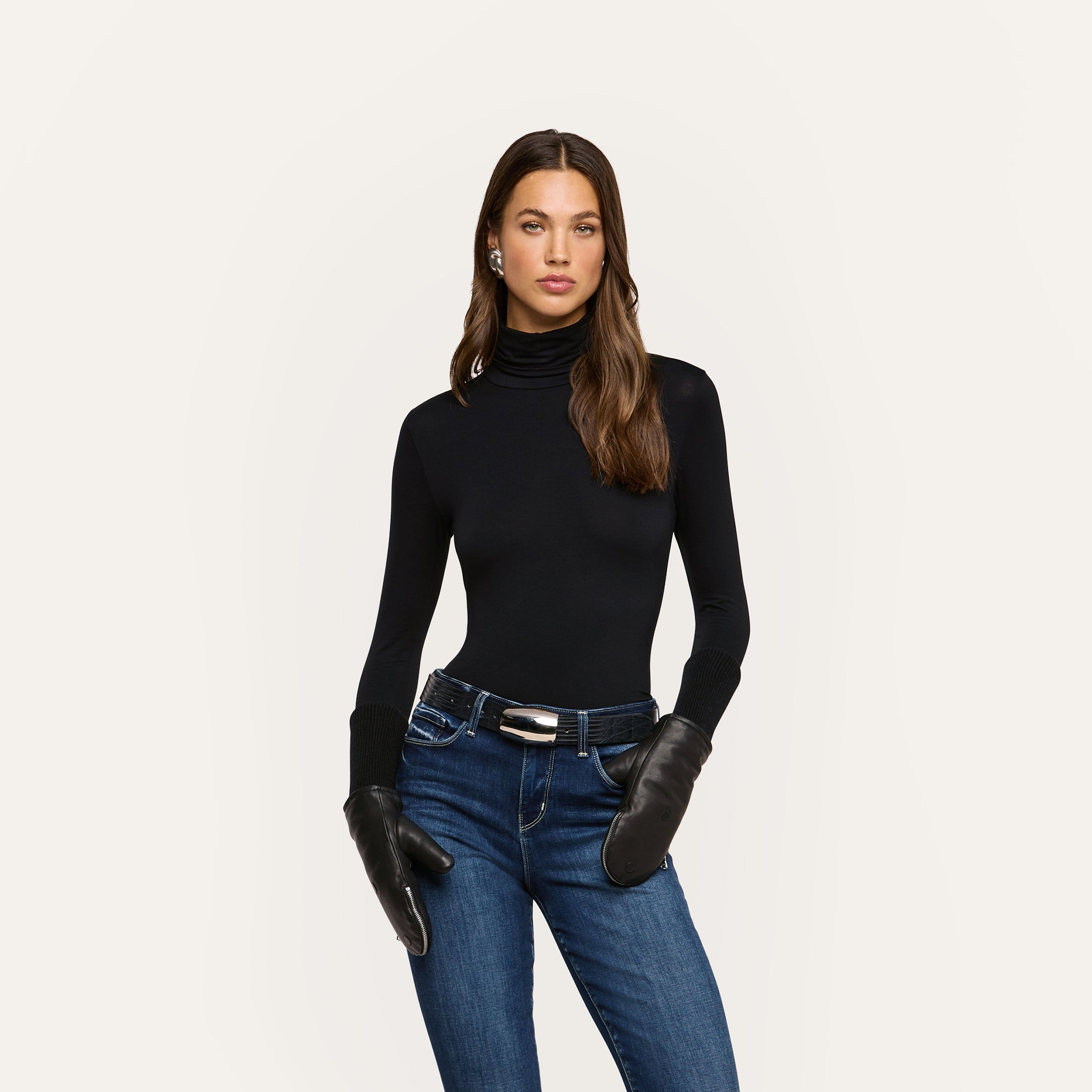 A woman with long brown hair wearing a black turtleneck, blue jeans, a belt, and earrings stands confidently against a plain light background. She completes her elegant ensemble with Tempo Mittens, adding a touch of sophistication to her look.